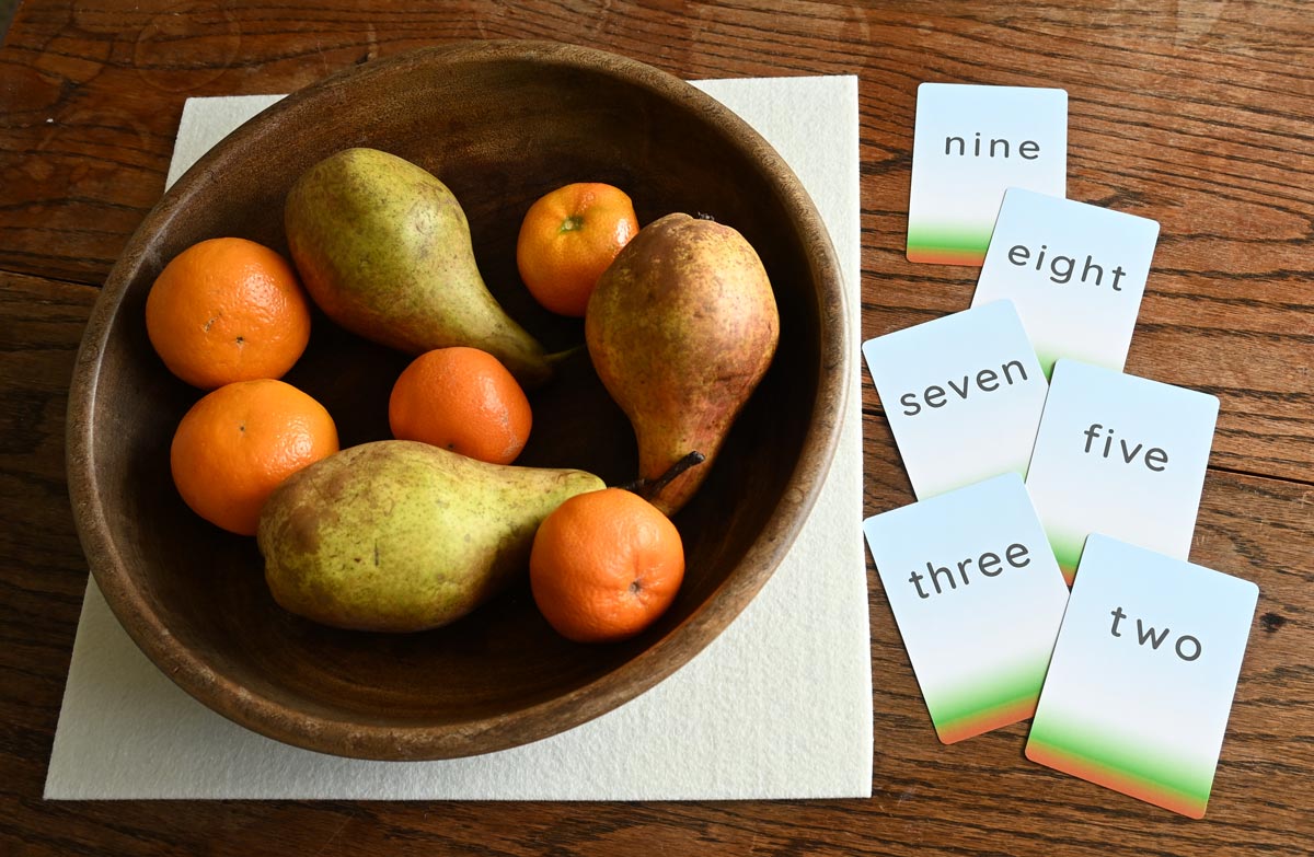 Three pears and five small oranges (mandarins) are in a bowl. Alongside is a selection of cards showing the words 'two', 'three', 'five', 'seven', 'eight', 'nine'